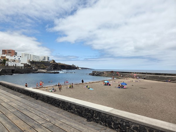 Ein kleiner Strand in Bajamar mit ruhigem Wasser, einer Steinmauer und einigen Badegästen unter leicht bewölktem Himmel.