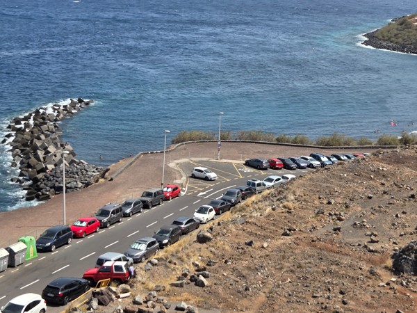 Blick von oben auf die Küstenstraße bei Playa de la Nea mit parkenden Autos, Wellenbrecher und blauem Meer.