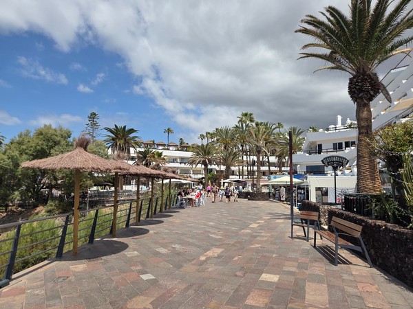 Breite Promenade in Costa Adeje mit Palmen, Sitzbänken, Restaurants und dramatischen Wolken über dem Urlaubsort.