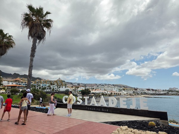 Promenade in Costa Adeje mit großem Schriftzug, Palmen, Strand und Hotels unter dramatischen Wolken.