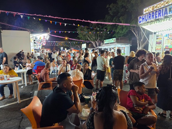 Belebtes Straßenfest bei Nacht mit vielen Menschen, bunten Girlanden und beleuchteten Essensständen wie einer Churrería.