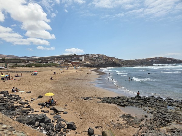 Ein breiter Sandstrand mit Badenden, Felsen und Atlantikwellen unter blauem Himmel an der Küste.