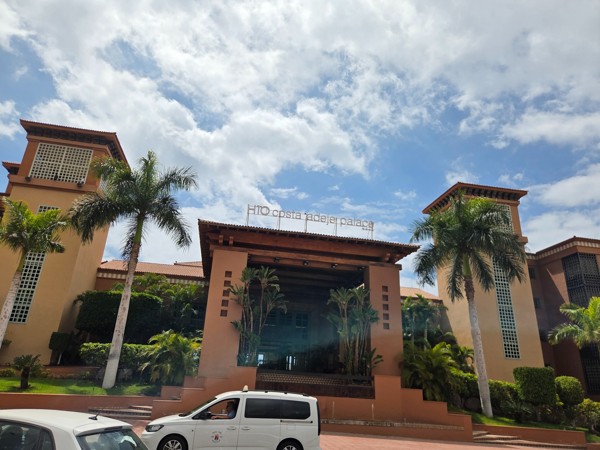 Eingangsbereich des Hotels H10 Costa Adeje Palace mit Palmen, markanter Architektur und parkenden Fahrzeugen davor.
