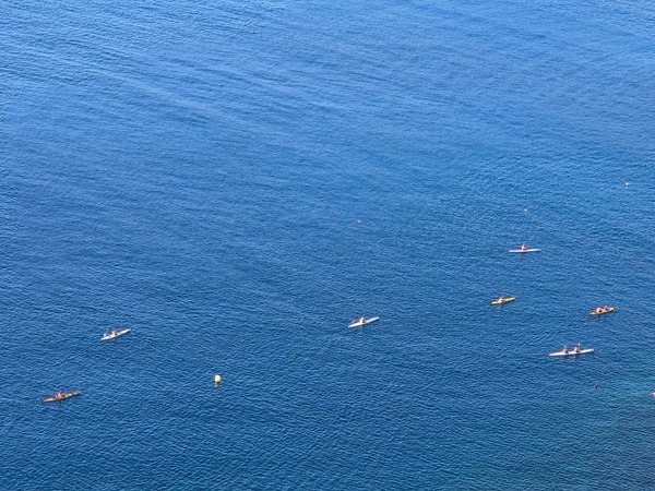 Mehrere Kajaks und Paddler fahren auf ruhigem blauem Meer, aus erhöhter Perspektive fotografiert.