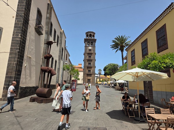 Eine sonnige Altstadtstraße in La Laguna mit historischem Turm, Cafétischen und spazierenden Menschen.