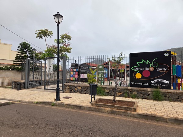Eingang zum Mercadillo de Tegueste mit Informationsschildern, Zaun und bewölktem Himmel im Hintergrund.