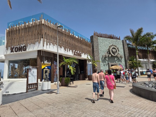 Außenansicht des Monkey Beach Club mit modernem Gebäude, Fußgängern, Palmen und sonnigem Himmel in Costa Adeje.