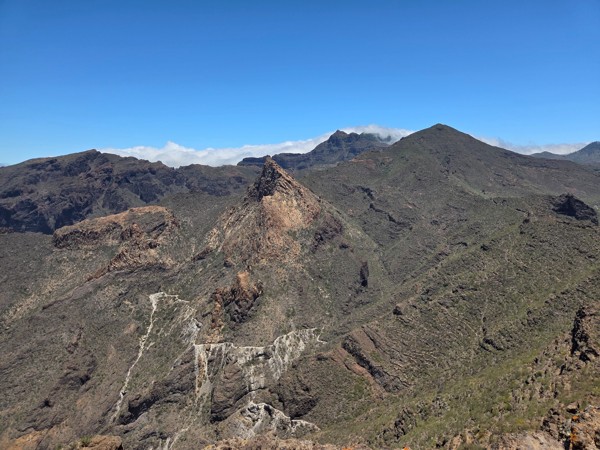 Berglandschaft der Montaña de Guama mit felsigen Gipfeln, kurviger Straße und wolkenbedeckten Höhen unter blauem Himmel.