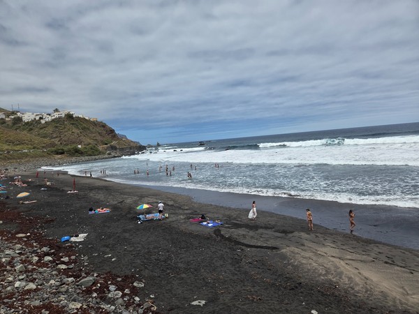 Blick auf den schwarzen Sandstrand von Almáciga mit Badenden, Wellen und den Häusern am Hang unter bewölktem Himmel.