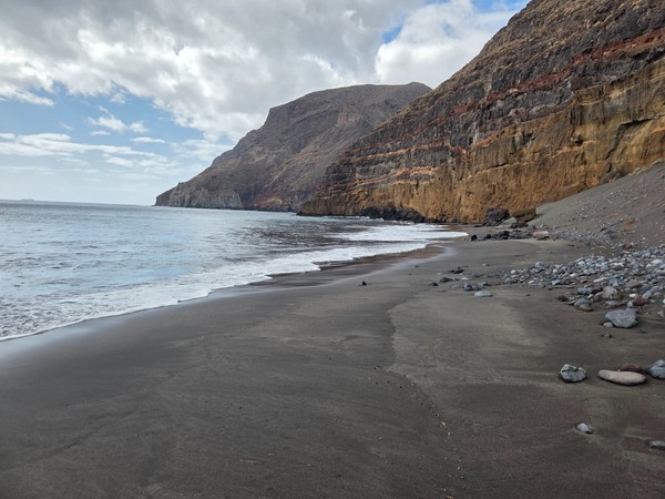 Ruhiger schwarzer Sandstrand von Antequera mit steilen Felswänden, sanften Wellen und einer abgelegenen Küstenlandschaft.
