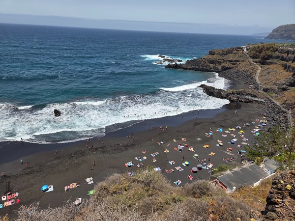 Panoramablick auf den schwarzen Strand Bollullo mit vielen Badegästen, kräftiger Brandung und tiefblauem Atlantik.