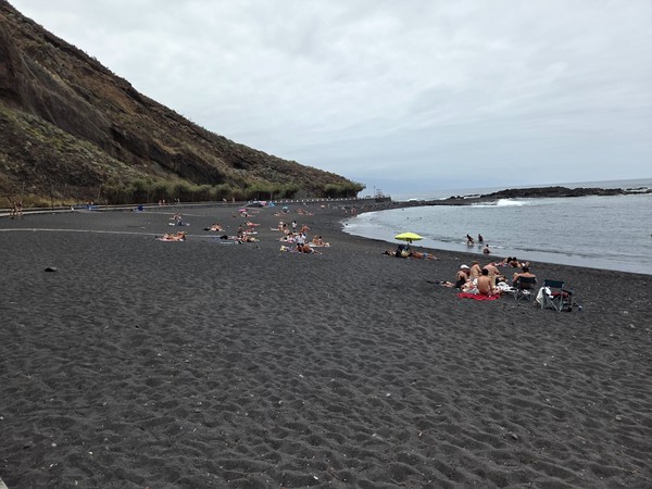 Menschen entspannen an der Playa de la Arena neben ruhigem Wasser und dunklem Vulkansand.