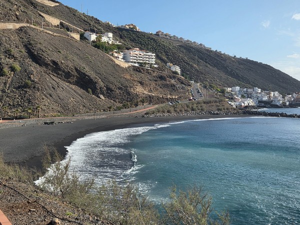 Kleine Bucht an der Playa de la Nea mit dunklem Sand, türkisfarbenem Wasser und Häusern am steilen Hang.
