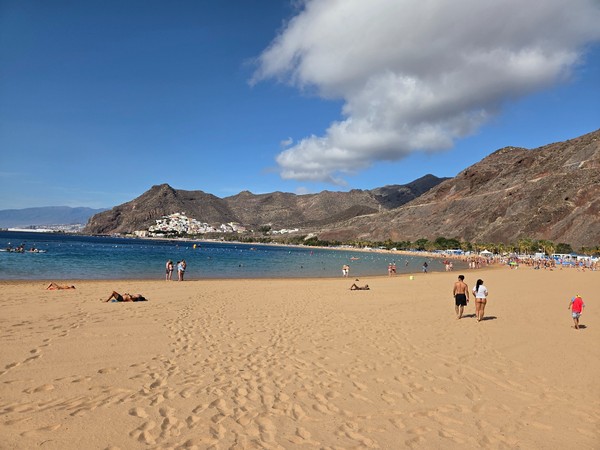 Weitläufiger goldener Sandstrand Las Teresitas mit ruhigem Wasser, Bergen im Hintergrund und Spaziergängern am Ufer.