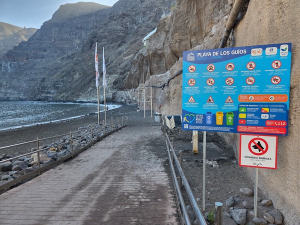 Zugang zur Playa de Los Guíos mit Holzweg, Hinweisschildern und steilen Felswänden direkt am schwarzen Strand.
