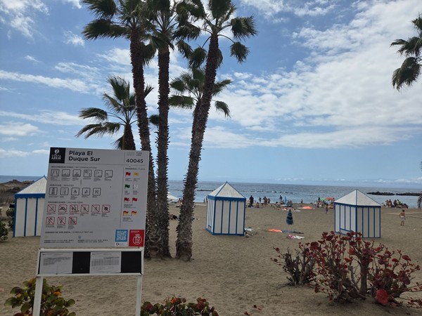 Ein Schild von Playa El Duque Sur steht zwischen Palmen und kleinen Strandkabinen mit Blick auf das Meer.