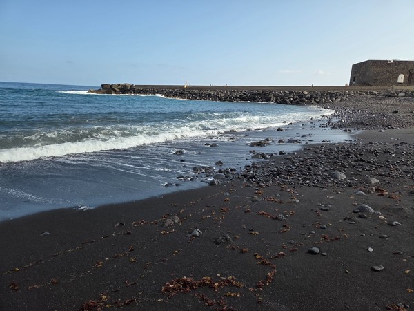 Sanfte Wellen rollen an den dunklen Sand der Playa El Jardín, daneben liegt eine lange Mole aus Steinen.