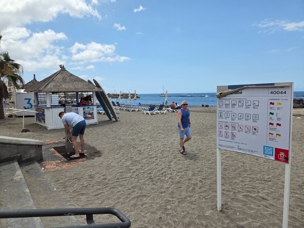 Am Playa Fañabé sieht man Strandliegen, einen kleinen Kiosk und ein Hinweisschild direkt am sandigen Zugang zum Meer.