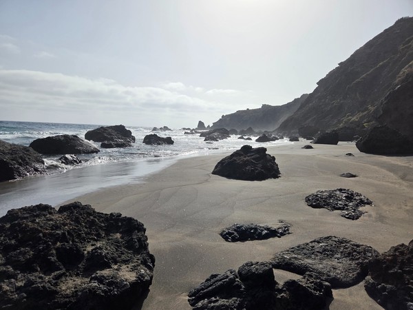 Abgelegener schwarzer Sandstrand mit großen Felsen, Brandung und hohen Küstenhängen im Gegenlicht.