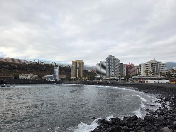 Küste von Puerto de la Cruz bei Playa Martiánez mit dunklem Ufer, Stadtgebäuden und wolkenverhangenem Himmel.