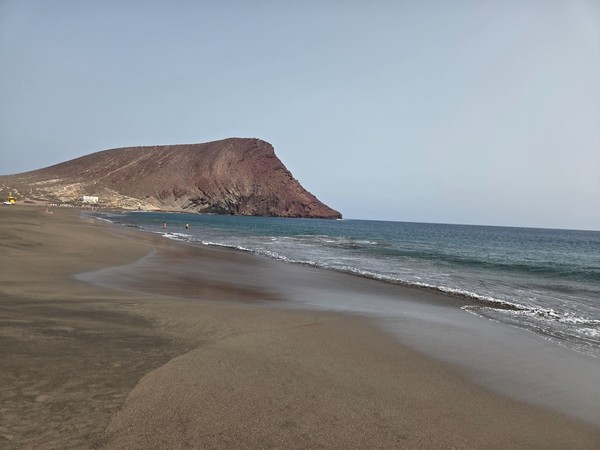 Breiter Strand La Tejita mit glattem Sand, ruhigem Meer und dem markanten roten Berg Montaña Roja im Hintergrund.