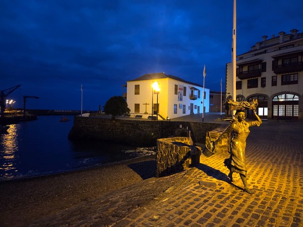 Nächtliche Szene im Hafen von Puerto de la Cruz mit beleuchteter Statue, historischem Gebäude und dunklem Himmel.