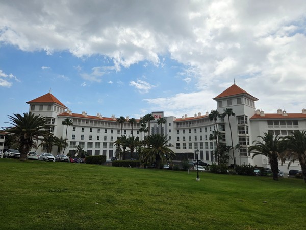 Das weiße RIU-Hotel in Costa Adeje steht vor einer gepflegten Rasenfläche mit Palmen und bewölktem Himmel.