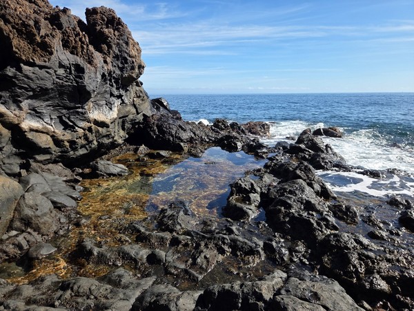 Schwarze vulkanische Felsen und kleine Wasserbecken liegen direkt am Meer unter einem klaren blauen Himmel.