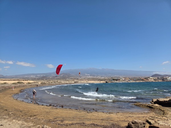 Ein Kitesurfer und mehrere Spaziergänger befinden sich an einer windigen Küste mit Blick auf Meer, Strand und Berge im Hintergrund.